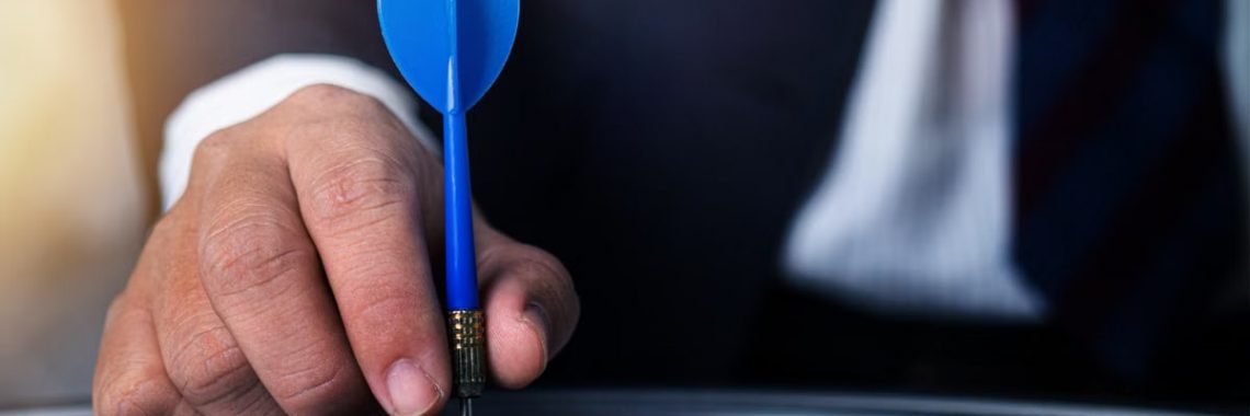 Close-up of a man's hand holding a blue dart on a bullseye, symbolizing precision and focus.